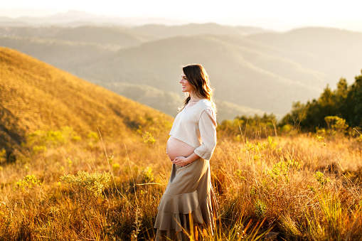 Curitiba, Brazil - August 11, 2021: Portrait of a beautiful pregnant woman showing her naked belly in the hills at sunset.