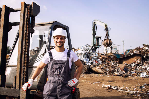 istock Portrait of worker standing by forklift while hydraulic industrial machine with claw attachment working with scrap metal parts in junk yard. 1333611569