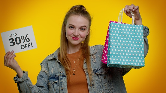 Cheerful teenager girl showing shopping bags and Up To 30 Percent Off inscriptions banner text. Black Friday. Good holiday discounts, low prices for shopping. Young woman posing on yellow background