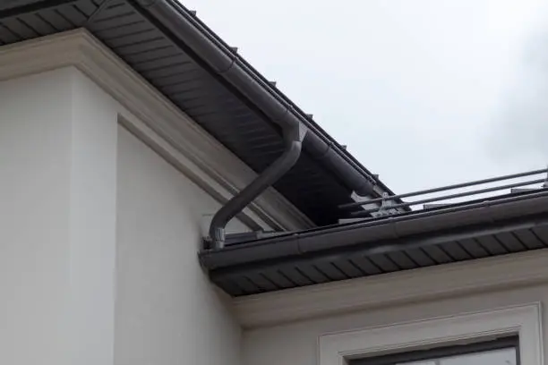 Photo of Corner of house with new gray metal tile roof and rain gutter. Metallic Guttering System, Guttering and Drainage Pipe Exterior