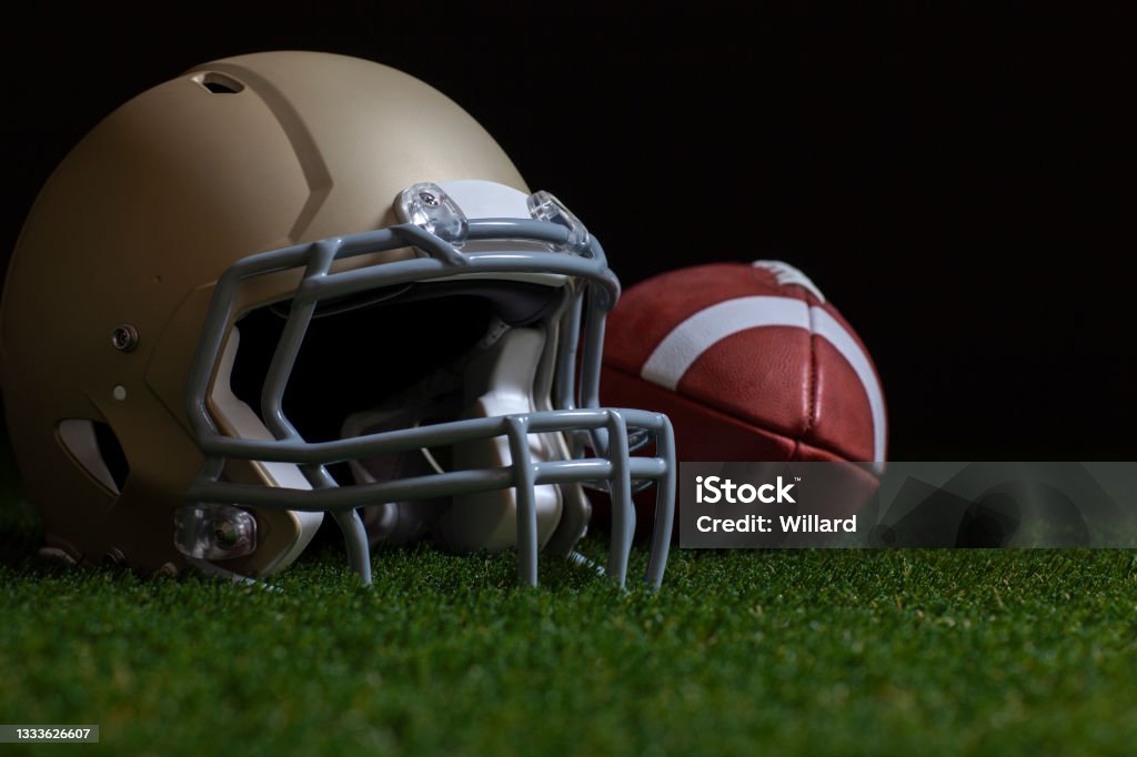 Low angle selective focus of football and gold helmet on grass with dark background College American Football Stock Photo