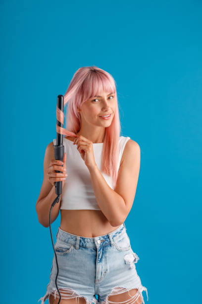 istock Cute woman smiling at camera while curling pink long straight hair using curling iron, standing isolated over blue studio background 1333633895