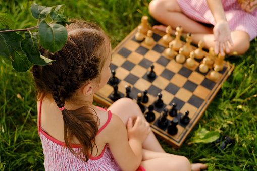 Two little girls are playing chess on a wooden chessboard outdoors in nature. One of the girls teaches the game and the rules of another child. Close-up. Rear view