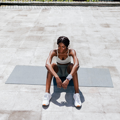 Beautiful African American woman sitting on grey mat with arms on knees after working out