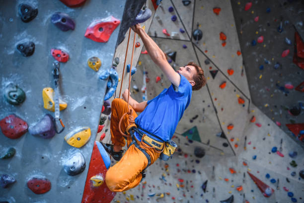 Personal perspective of sportsman in early 20s concentrating on hold route as he scales wall in modern sport climbing center.