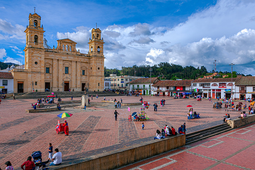 Chiquinquirá, Colombia - November 26, 2017: It is Sunday afternoon on the Plaza Mayor in the Andes town of Chiquinquirá in the Department of Boyacá, in the South American Country of Colombia. Many local residents and Colombian tourists are seen enjoying the ambience of the town square. Vendors sell toys and snacks to visitors. In the background is the 19th Century Basilica of Our Lady of the Rosary. It houses the famous painting of Our Lady of The Rosary, also referred to as the Virgin of Chiquinquirá, the Patron Saint of Colombia. It is designed in the Neo-classical style of architecture and is a destination of Catholic pilgrimage in South America. The altitude at street level is about 8400 feet above mean sea level. Photo shot in the late afternoon sunlight; horizontal format.