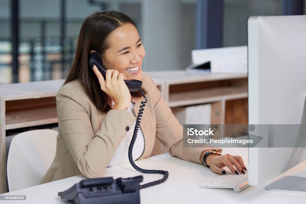 Shot of a young woman using a telephone and computer in a modern office You've called the right company Using Phone Stock Photo
