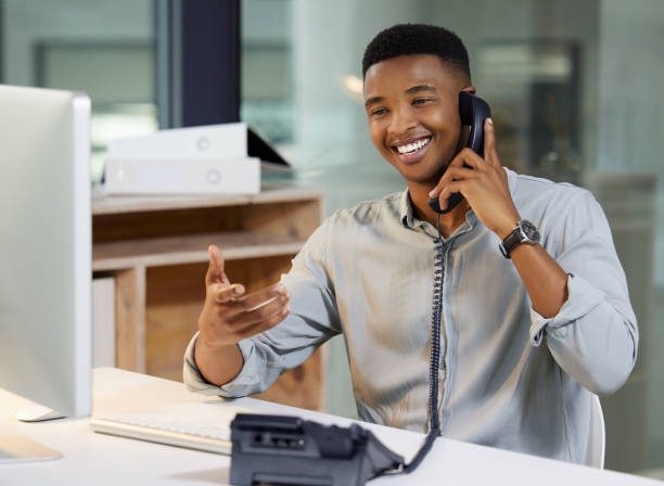 Shot of a young man using a telephone and computer in a modern office That's what we love to hear landline phone stock pictures, royalty-free photos & images