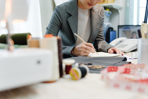 Close-up Hands of female fashion designer or tailor while startup drawing on daily book in cutting and design studio of dressmaker. Very shallow focus point at her hand while note.