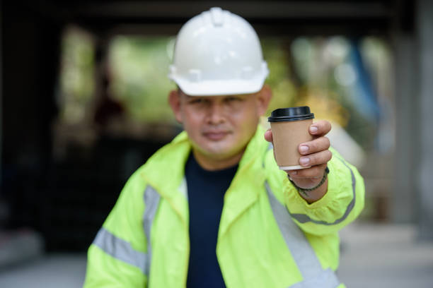 istock Construction engineer in reflective unifrom holds a cup hot coffee and hand to camera before work begins. Selection focuses on the cup of coffee. 1333691221