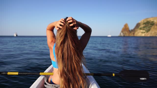 Young attractive brunette woman with long hair in white swimsuit, swimming on kayak around volcanic rocks, like in Iceland. Back view. Summer holiday vacation and travel concept
