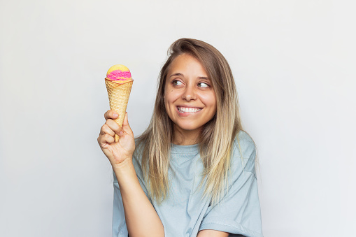 A young caucasian excited pretty blonde woman in a gray t-shirt smiles looking at the fruit ice-cream sorbet in a waffle cone biting her lip on a white background. Dieting, temptation, seduction