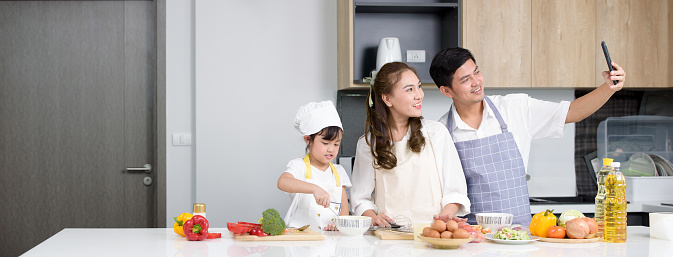 Asian family blogger happiness selfie and taking picture by smartphone while preparing salad in the kitchen for teaching daughter to cook homemade food on the day at home.family.