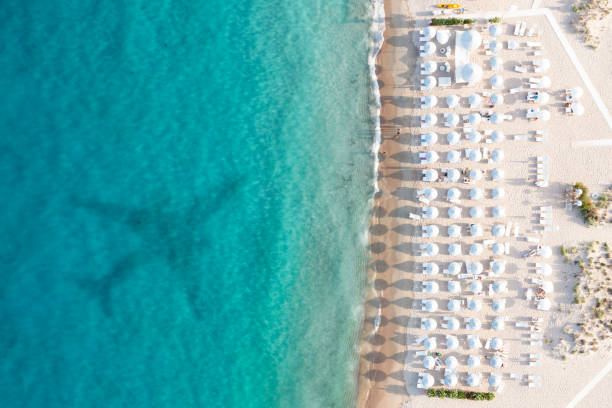 Stunning aerial view of a white sand beach bathed by a turquoise water with some white beach umbrellas and the shadow of an airplane landing in Sardinia. Cala di volpe beach, Sardinia, Italy. Stunning aerial view of a white sand beach bathed by a turquoise water with some white beach umbrellas and the shadow of an airplane landing in Sardinia. Cala di volpe beach, Sardinia, Italy. Cala Di Volpe stock pictures, royalty-free photos & images