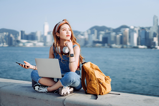Thoughtful young Asian woman sitting cross-legged by the promenade, against urban city skyline. She is wearing headphones around neck, using smartphone and working on laptop. Teenage lifestyle and technology