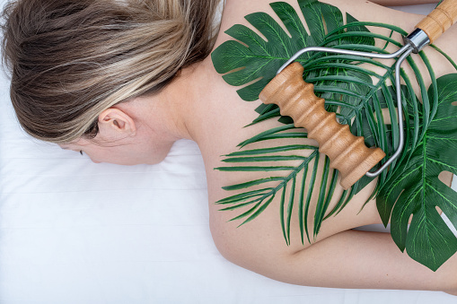 Madero therapy, relaxing massage - woman having spa back massage using a wooden roller massager lying on green leaves on her back, close-up, top view. Body care concept