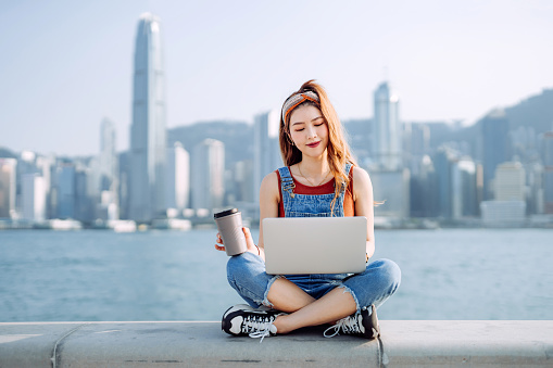 Beautiful young Asian woman sitting by the promenade, against urban city skyline. She is using laptop on lap, drinking with a reusable coffee cup. Teenage lifestyle and technology