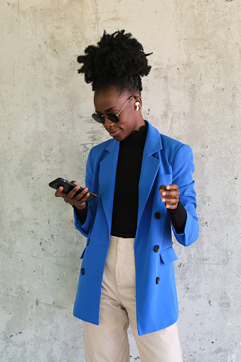 Portrait of smiling young african woman wearing wireless earbuds, selecting the music with her smartphone, wearing blue jacket suit and sunglasses over a concrete wall.