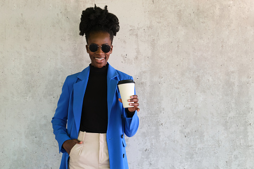 Portrait of smiling young african woman wearing a blue jacket suit, sunglasses and holiding a coffee over a concrete wall.