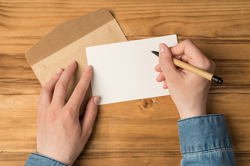 First person top view photo of hands holding pen over white card and craft paper envelope on isolated wooden table background with blank space