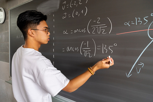 Teen asian high school boy student solving maths exercise on blackboard with chalk. Education concept.
