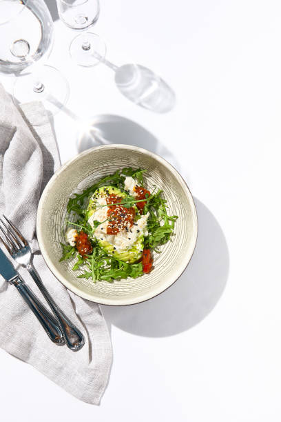 istock Avocado and stracciatella cheese salad. Fancy dinning with Italian salad bowl on white table with simple contemporary decor. Sunlight and harsh shadow still life. Avocado, cheese and tomato salad. 1333733905