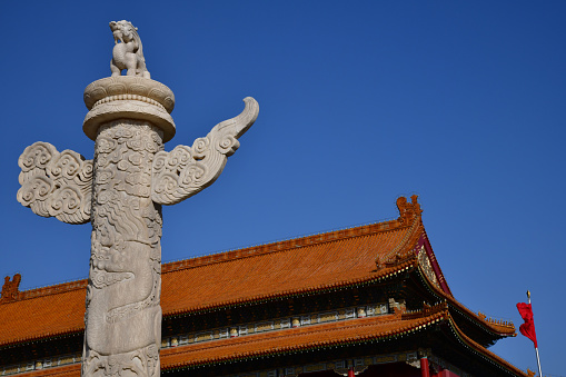 Beijing, China - October 2019; Low angle close up view of a marble huabiao column in front of  one of the iconic red roofs of the Forbidden city in Beijing against a blue sky; on top of the column the mythical creature denglong, one of the ‘nine children of the dragon’
