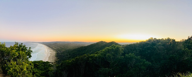 Horizontal panorama of ocean waves breaking onto the sand at Tallow Beach, rainforest covered hills and coastal scrub forest of the Arakwal National Park at sunset in Winter.
