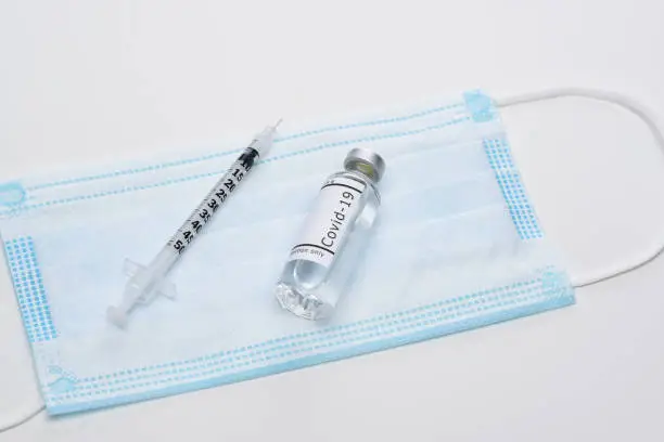 Covid-19 Concept. A vial of vaccine surrounded and syringe on a surgical mask, on white. The bottle has no label.