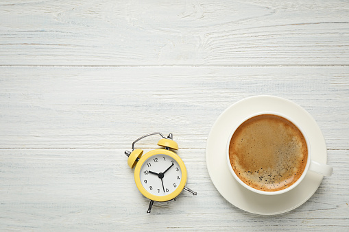 Cup of morning coffee and alarm clock on white wooden table, flat lay. Space for text