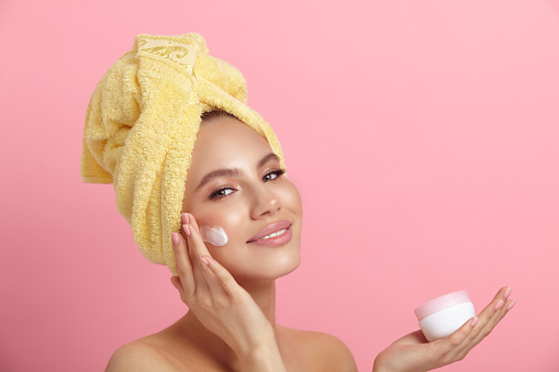 Perfect skin secret. Smiling lady with terry towel on head applies cream on face showing small jar on pink background closeup