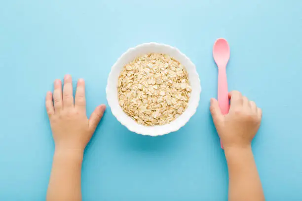 Baby hand holding pink plastic spoon. White bowl with dry rolled oat on light blue table background. Pastel color. Closeup. Point of view shot. Children healthy food. Top down view.