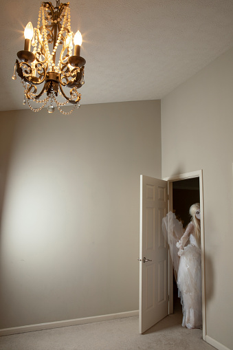 A creepy ghost bride figure peeking in a door of a big room with a chandelier.