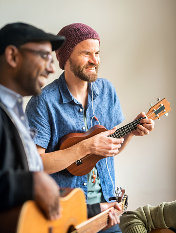 Smiling young man playing a ukulele during a band rehearsal with friends in a home recording studio