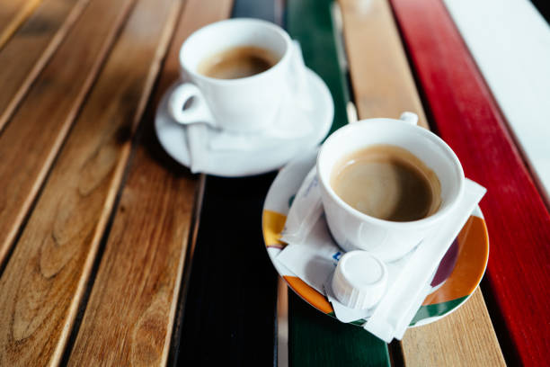Selective focus color image depicting a cup of fresh coffee on a cafe table outdoors. The wooden table is painted in colorful stripes.