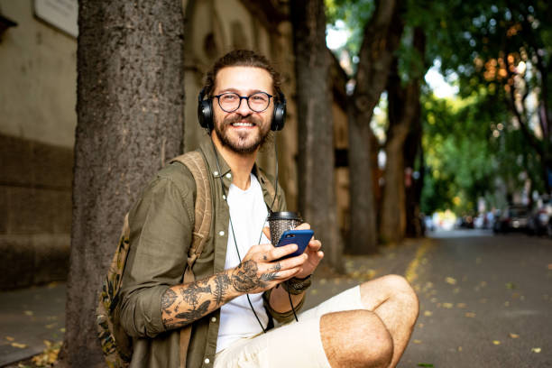 Young modern man at the city street enjoying the cup of coffee while listening music on headphones, using mobile phone Young modern man at the city street enjoying the cup of coffee while listening music on headphones, using mobile phone hipster person stock pictures, royalty-free photos & images