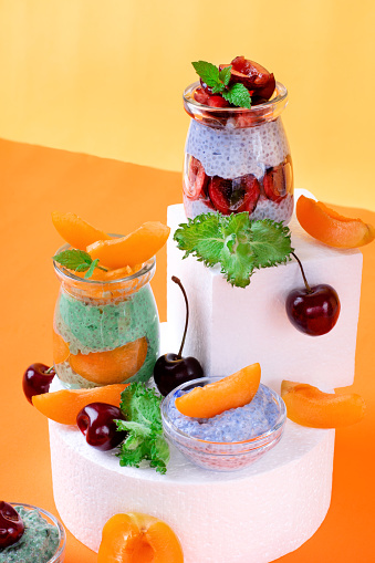 Chia pudding with spirulina and matcha tea powders topped with cherry and apricot in glass jars on white podiums against the colored background. Healthy dessert