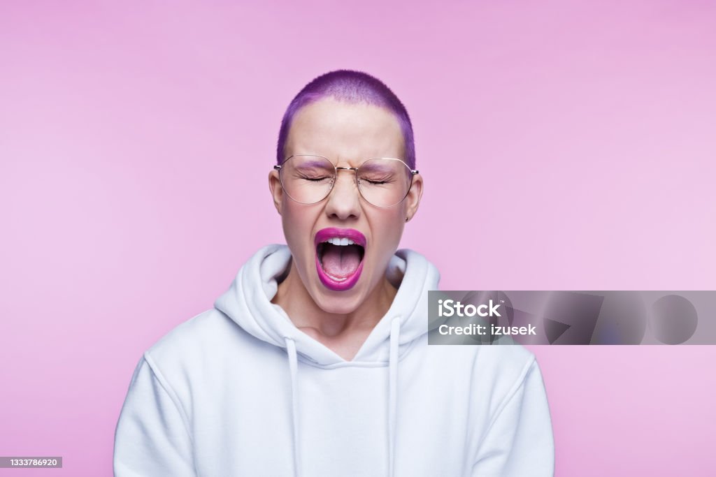 Angry young woman with short purple hair Headshot of displeased young woman wearing white hoodie, screaming at camera with eyes closed. Studio shot on pink background. Shouting Stock Photo