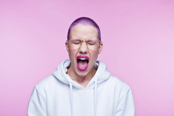 Angry young woman with short purple hair Headshot of displeased young woman wearing white hoodie, screaming at camera with eyes closed. Studio shot on pink background. rolling eyes irritation displeased facial expression stock pictures, royalty-free photos & images