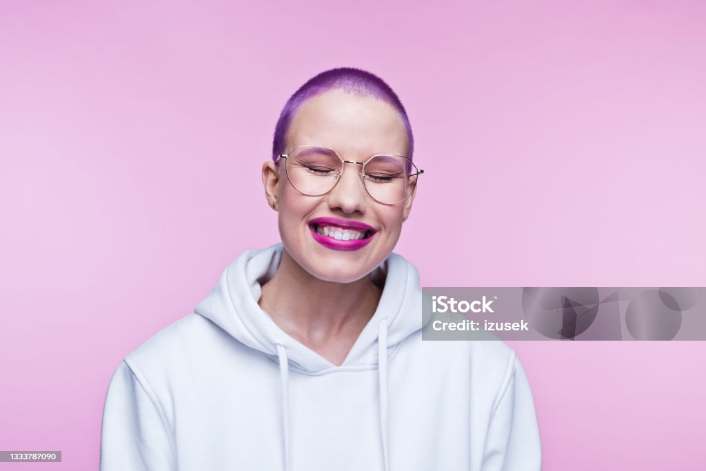 Cheerful young woman with short purple hair Headshot of happy young woman wearing white hoodie, laughing with eyes closed. Studio shot on pink background. One Woman Only Stock Photo