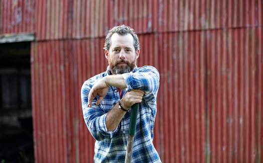 A mature man in his 40s with a thick beard, wearing a plaid shirt, working on a small family owned farm, standing in front of a red barn. He is leaning on the handle of a hoe, looking at the camera with a serious expression.