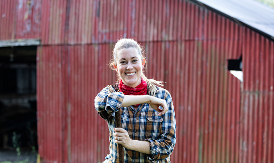 Headshot of an Hispanic woman in her 30s, wearing a plaid shirt, working on a small family owned farm, standing in front of a red barn. She is leaning on the handle of a hoe, looking at the camera, smiling.