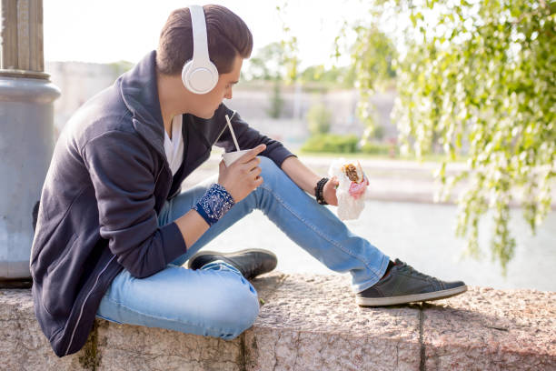 Young Man sitting in the deck by the river, Stylish guy sits on the quay on the background of the river and the bridge. He wears blue jeans. Male looks up. Outdoors. Horizontal. wood and wire fence designs pictures stock pictures, royalty-free photos & images