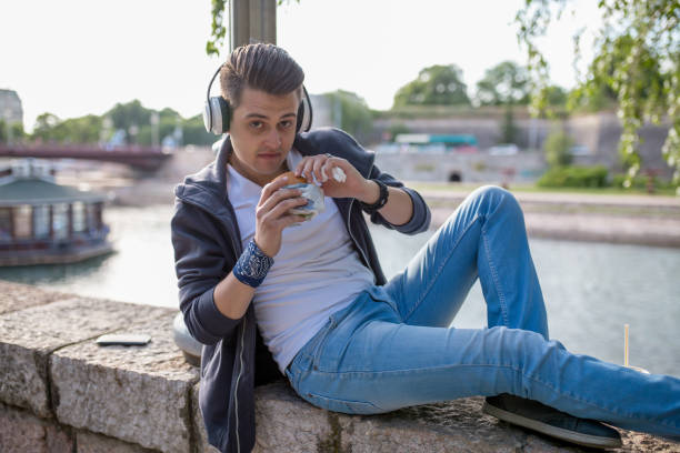 Young Man sitting in the deck by the river, Stylish guy sits on the quay on the background of the river and the bridge. He wears blue jeans. Male looks up. Outdoors. Horizontal. wood and wire fence designs pictures stock pictures, royalty-free photos & images