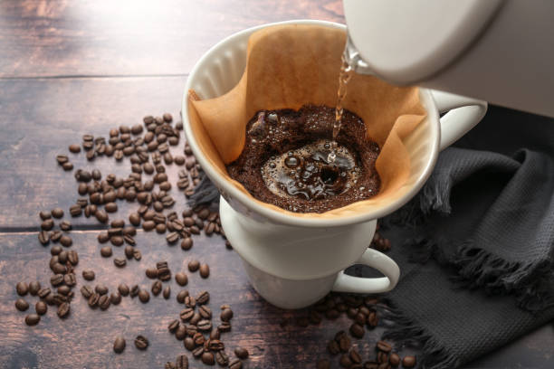 Brewed coffee, pouring hot water on ground coffee in a filter on a mug for a stimulating drink, rustic wooden table with some beans, copy space, selected focus, narrow depth of field stock photo