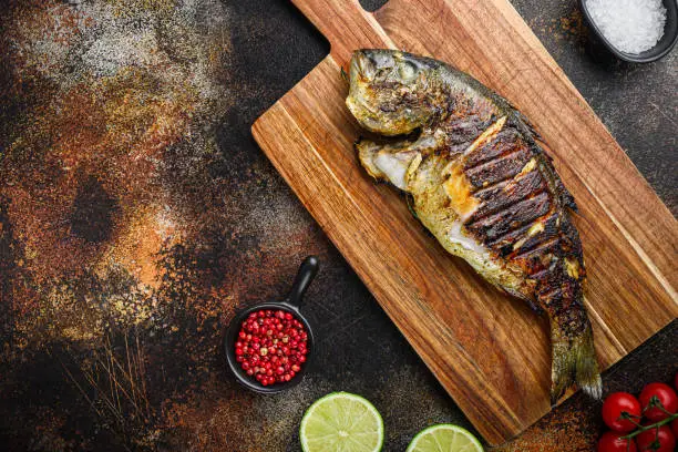 Grilled sea bream or dorado raw fish on chopping  board with ingredients over old metall dark background, top view with space for text