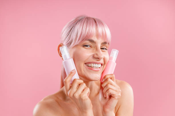 jeune femme heureuse aux cheveux roses tenant deux bouteilles en plastique avec des produits de beauté près de son visage, posant isolée sur fond rose - spay photos et images de collection