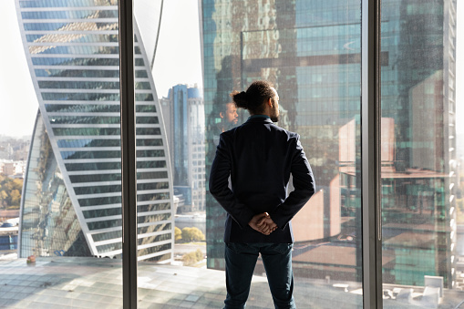 Back rear view confident successful young african american businessman in suit standing near huge panoramic window with skyscrapers view, thinking oof problem solution, planning development strategy.