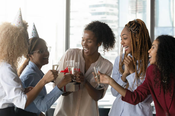 Happy african american colleagues celebrating birthday. Overjoyed emotional sincere millennial generation african american multiethnic business women wearing paper party hats, celebrating birthday or working anniversary, enjoying hangout in workplace. business party stock pictures, royalty-free photos & images