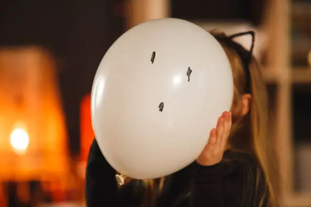 Portrait of little girl playfully holding a white balloon in front of her face, looking towards the camera, as a Halloween mask. She drew a ghost face on it.